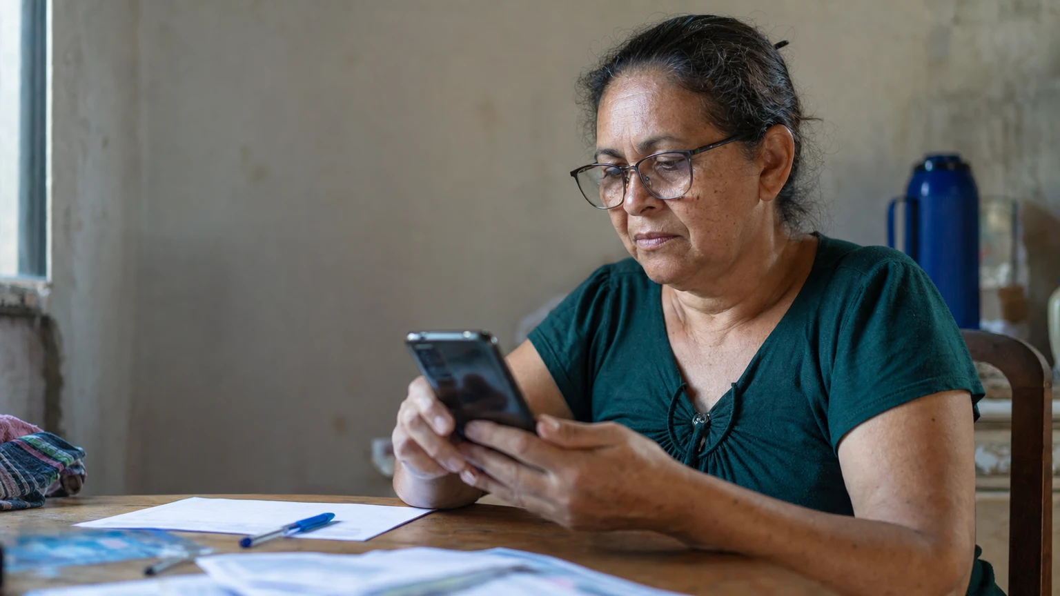 Pessoa em casa usando o celular para consultar os canais oficiais do INSS (Meu INSS e telefone 135), com documentos ao lado na mesa.